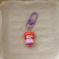 Close-up of cute cartoon girl keychain with orange hair and purple dress, bag charm. - Purple