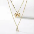 Primo piano della collana dorata a doppio strato con fiocco e ciondolo a lettera brillante. - Stile a Doppio Strato con Lettera Iniziale A e Fiocco / Oro