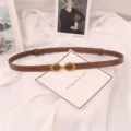 Cinturón fino marrón minimalista para mujer con hebilla dorada, detalle elegante sobre tela blanca. - Caramelo