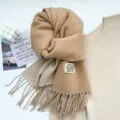 Close-up detail of soft camel winter tassel scarf showing fabric texture and woven brand label. - Beige