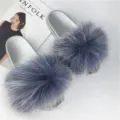 Detail view of stylish silver PVC slides featuring large fluffy grey raccoon fur pom-poms - Cyan
