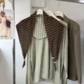 Detail shot of a chic striped triangular scarf layered with knit top on hanger in a stylish showroom setup. - 06_Coffee Striped