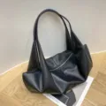 Detailed close-up shot of a black minimalist slouchy shoulder tote bag on wooden floor. - Solid Black