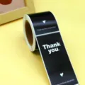 Roll of black thank you sealing stickers detail showing clear white text and heart design. - Black