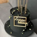 Black birthday cake with gold candles and acrylic happy birthday topper for party decor. - Gold