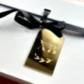 Close-up detail of gold mirror acrylic pendant tag with black ribbon on white box packaging. - Gold XB Pendant