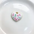 Close-up detail of the white enamel heart pendant with colorful love text and gold trim. - LOVE IS ALL Style (C01-8)