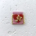 Close-up detail of handmade pink woven fabric charm with red heart and gold Fe letter. - Pink