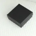 Close-up van een zwarte vierkante sieradengeschenkdoos met diamantpatroon op witte stof. - Zwarte Geschenkdoos