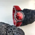 Detalle reloj cuarzo esfera roja, índices dorados, caja negra, correa elástica extensible. - Rojo