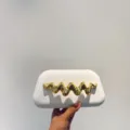 Detail shot of a white matte clutch bag featuring an elegant gold wavy metal decoration. - White