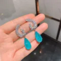Gedetailleerde close-up van oorknopjes met fonkelende zirkonia vleugels en blauwe peer edelsteen. - Paraiba Blauw Oorknopjes