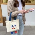 Cute canvas lunch tote bag with adorable embroidered face and dangling feet design for daily use. - Freckle Smiley Face Little Legs Bento White
