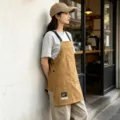 Khaki canvas work apron with cross-back straps, practical pockets, stylish barista uniform. - Webbing Utility Style / Camel