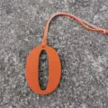 Orange leather number 0 charm detail shot, fine hanging strap, textured surface closeup view. - Orange