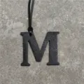 Detailed close-up of a black leather letter M bag charm with strap on textured surface. - Black
