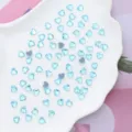 Close up of light blue heart-shaped crystal rhinestones scattered on a white shell dish for nail art - 6x6mm 100 Pieces/Pack / Lake Blue