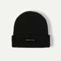 Detailed shot of a black winter knit beanie hat, featuring a woven label with text accent. - One Size / Black