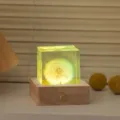 Close up of the glowing dandelion resin cube night light on wood base in warm bedroom setting. - 5cm Resin Cube + Touch Charging Base (Colorful Dandelion)