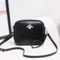 Detail view of the black leather crossbody bag featuring a delicate gold bee and pearl ornament. - Black