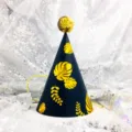 Close-up detail of a black and gold tropical leaf birthday party hat with a shiny pompom. - Blue Background Leaves (Sequin Ball)