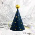 Close up detail view of a dark blue children birthday party cone hat with gold stars and pompom. - Blue Background Stars (Sequin Ball)