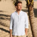 Detailed view of white linen long sleeve mandarin collar shirt worn on beach scene. - S / Tops / White