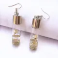 Close up of clear glass bottle earrings filled with tiny white flowers, liquid and gold glitter. - White