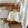 Cream knitted baby cardigan with lace collar, hanging on wooden background detail view. - 66cm / Apricot