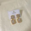 Eleganti orecchini pendenti geometrici in zircone dorati su cartesio di presentazione. - Quadrato Bianco