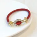Dettaglio di un bracciale in pelle intrecciata rossa con zirconi e finiture in metallo dorato di lusso. - Rosso