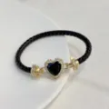 Primo piano del bracciale in pelle intrecciata nera con chiusura in zircone a cuore dorato. - Nero