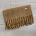 Detailaufnahme des braunen Winterschals mit dicken Fransen und kleinem Metalllogo. - 40x220cm / Kastanienbraun