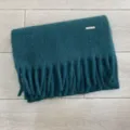 Detailansicht des dunkelgrünen Winter Fransenschals mit feinem Metalllogo aus weichem Stoff. - 40x220cm / Dunkelgrün