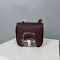 Detail view of the burgundy leather saddle crossbody bag, showing the metal clasp. - 19 / Wine Coffee