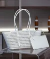 Detail shot of white perforated leather tote bag with matching pouch on clear glass table surface - L31xW15xH21cm with Gold Buckle / White