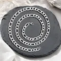 Gros plan sur un collier à chaîne Figaro en argent poli sur une dalle de pierre noire texturée. - 60CM / Chaîne Figaro (XL2002-23) / 2.0NK3-1 / Acier