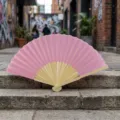 Elegant pink silk folding fan with bamboo ribs, displayed on outdoor stone steps. - Style Q No.10 - Smooth Colored Fan (with Border) / Magenta Pink