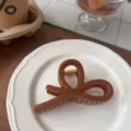 Close-up of a brown resin bow hair claw clip placed on a white ceramic plate, detail view. - Single Pack (Approx 11cm) / Caramel Brown - Bow Knot Style