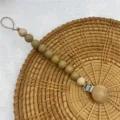 Silicone pacifier clip detail closeup showing wooden beads and metal clasp on rattan background. - Oatmeal