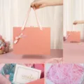 Boîte cadeau rose à nœud avec poignée, raphia et carte pour emballer parfum et bonbons. - Boîte cadeau rose avec nœud à bordure dorée + raphia + carte