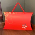 Red pillow-shaped gift box with rope handle and gold floral detail for packaging presents. - Red Ingot Box