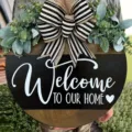 Close-up of wooden welcome door sign with striped bow and green eucalyptus leaves - Bless This Home Style / White with Heart