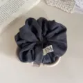 Close-up van een donkergrijze stoffen haarscrunchie met een klein minimalistisch labeldetail. - Grijs