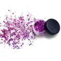 Detail view of purple cosmetic glitter sparkling powder in a small jar with loose flakes scattered. - Purple