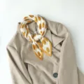 Detailed shot of pleated polka dot small square scarf tied neatly on neutral blazer collar. - Yellow