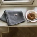 Detailansicht des gemütlichen grauen Strickschals auf der Fensterbank mit Lifestyle-Requisite. - 160x21cm / Grau