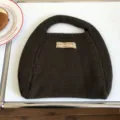 Nahaufnahme der braunen Strickhandtasche mit weicher Wollstruktur und einem Vintage-Stoffetikett. - 31x26cm / Dunkelbraun