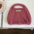 Detailansicht der rosa Strickhandtasche mit Etikett auf weissem Tisch im minimalistischen Stil. - 31x26cm / Rosa