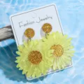 Nahaufnahme von hellgelben Acryl-Gänseblümchen-Ohrringen mit Glitzerdetail auf weißer Karte. - Gelbes Kleines Gänseblümchen